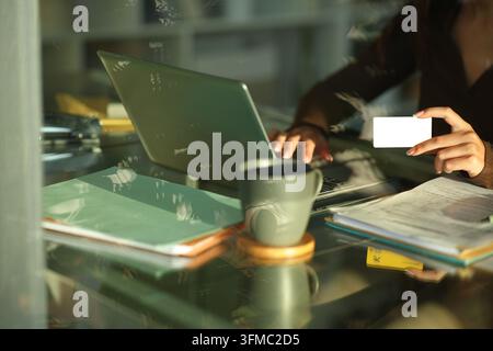 Nahaufnahmeporträt einer Fernarbeitshand, die einen Kreditkarten-Mockup hält, der Online-Einkäufe tätigt, während sie von zu Hause aus arbeitet, gesehen durch ein Fenster Stockfoto