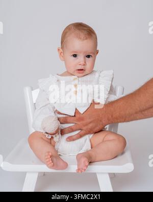 Baby sitzt auf einem Stuhl, vor weißem Innenhintergrund, mit weißem Hochstuhl, Hand hält das Baby, aus einer Nahaufnahme leicht über der Babyhöhe Stockfoto