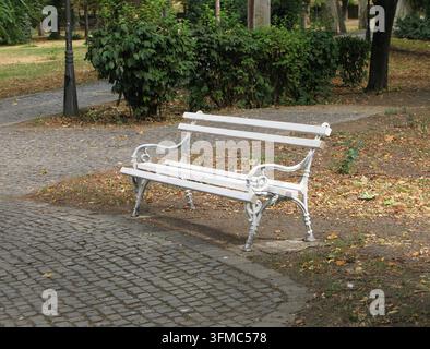 Verschnörkelte weiße Parkbank neben einem Kopfsteinpflasterweg in einem grünen öffentlichen Garten mit alten Bäumen Stockfoto