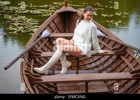 Junge Frau in weißem Pullover und kniehohen Stiefeln sitzt auf Holzboot in ruhigem See, umgeben von Seerosenblättern und üppiger Vegetation Stockfoto