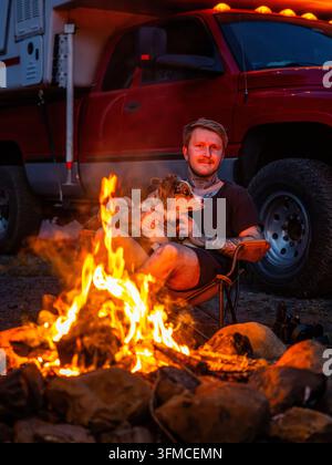 Junger tätowierter Mann hält einen Hund am Lagerfeuer bei Dämmerung Stockfoto