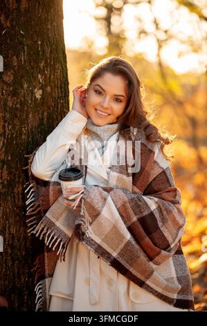 Lächelnde junge Frau in kuscheliger karierten Decke, die im Freien eine Kaffeetasse hält Stockfoto