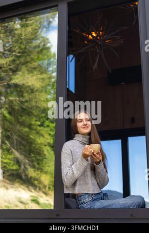 Junge Frau sitzt an einem offenen Fenster und hält ein warmes Getränk, gemütliches Lifestyle-Porträt in einem modernen Haus, umgeben von friedlichem grünen Wald. Stockfoto