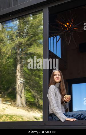 Junge Frau sitzt an einem offenen Fenster und hält ein warmes Getränk, friedliches Lifestyle-Porträt in einem modernen Waldhaus mit grüner Natur draußen. Stockfoto