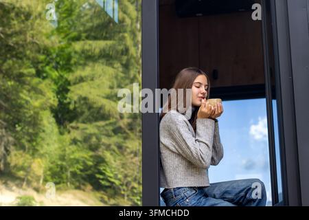 Junge Frau, die ihre Hände an einer Tasse wärmt, an einem offenen Fenster, ein gemütlicher Morgenmoment in einem modernen Waldhaus, umgeben von üppiger grüner Natur. Stockfoto