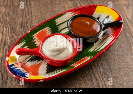 Tomato- und cremige Kräutersaucen in kleinen Schüsseln auf einem farbenfrohen usbekischen Ikat-Keramikteller, der auf einem Holztisch mit grünen Trauben platziert ist Stockfoto