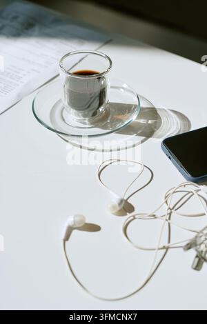 Gläserner Espresso-Becher mit Smartphone und Kopfhörern auf dem Schreibtisch Stockfoto
