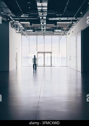 Vollständige Silhouette einer einsamen Person, die in einem hellen minimalistischen Galerieraum mit großen Glasfenstern steht, die auf das Wasser hinausgehen. Stockfoto