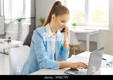 Junger Analyst, der am Bürotisch in sein Smartphone spricht und gleichzeitig auf dem Laptop tippt Stockfoto