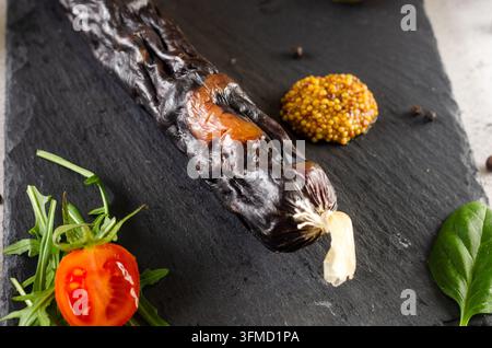 Ganze luftgetrocknete Wurst serviert auf einer schwarzen Schieferplatte mit Kirschtomaten, grünen Oliven, Senf aus der Mühle und Spinat Stockfoto