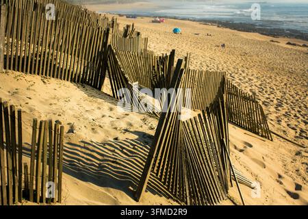 Verwitterte hölzerne Dünenzäune werfen gestreifte Schatten auf einen sandigen Strand mit Meereswellen im Hintergrund Stockfoto