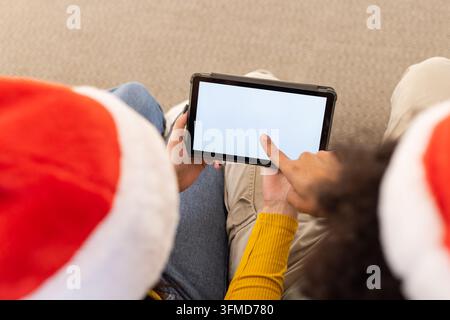 Afroamerikanisches Paar mit Weihnachtshüten zeigt auf ein schwarzes Tablet mit leerem Bildschirm auf dem Teppich Stockfoto