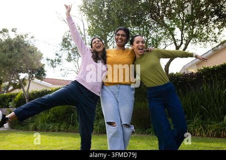 Drei Freundinnen lachen und geben sich auf einer grasbewachsenen Wiese die Arme, während sie Strickjacken und Jeans tragen Stockfoto