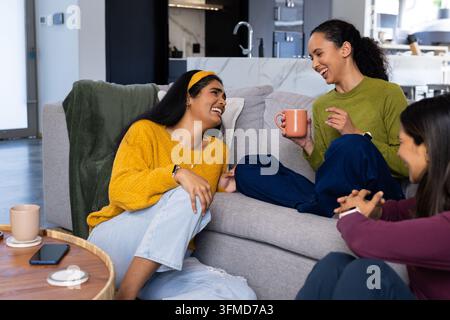 Verschiedene Freundinnen, die auf einem grauen Ecksofa zu Hause sitzen, lachen und eine Terrakotta-Tasse halten Stockfoto