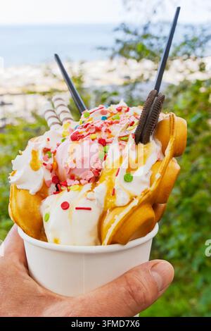 Nahaufnahme eines handgehaltenen Bubble-Waffle-Desserts mit rosa Eis, hellen Streuseln, Waffelrollen und Keksen in einer weißen Papptasse im Freien mit Grünpflanzen und Stockfoto