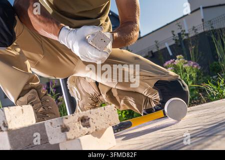 Nahaufnahme eines knienden Arbeiters, der einen Travertin-Fliesenstein hält, mit einem Gummihammer und einer Wasserwaage auf der Terrasse. Stockfoto