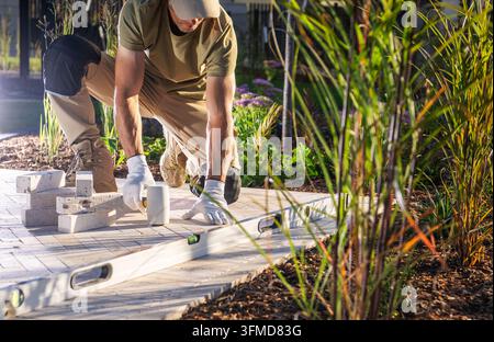 Ein kniender Arbeiter verwendet einen Gummihammer auf Travertin-Pflastersteinen neben Ziergräsern und einer langen Wasserwaage. Stockfoto