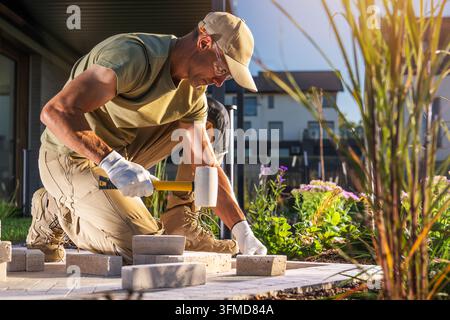 Ein Arbeiter kniet auf einer Gartenterrasse und verwendet einen Gummihammer beim Verlegen von Travertinplatten in der Nähe von Blumenbeeten. Stockfoto