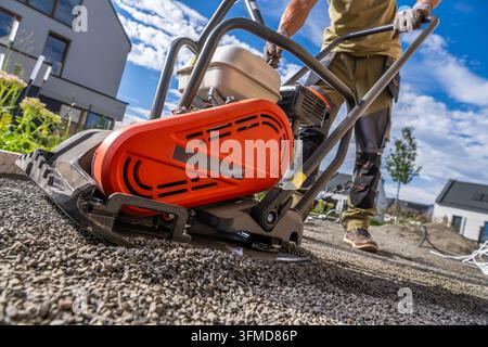 Niedriger Blick auf einen Arbeiter, der einen Vibrationsplattenverdichter auf einer Kiesunterlage in einem Wohngarten bedient. Stockfoto