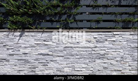 Detail eines Zauns aus weißer und grauer Kunststeinverkleidung mit schwarzen Metalllatten und Hecke oben. Hintergrund und Textur. Stockfoto