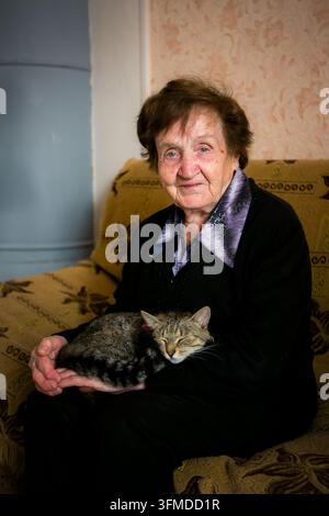 Porträt einer älteren Frau, die auf einem Sofa sitzt und eine schlafende getigerte Katze auf ihrem Schoß hält, zu Hause Stockfoto