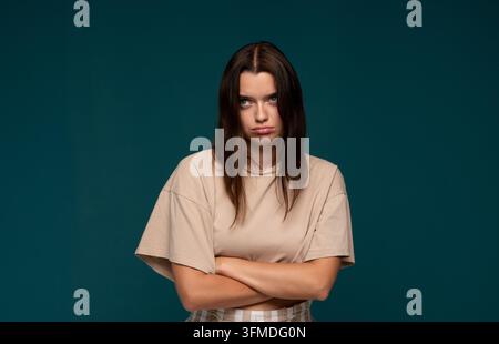 Verärgerte junge Frau, die mit gerunzelter Stirn und verschränkten Armen steht und Unzufriedenheit und Frustration auf einem blauen Hintergrund ausdrückt Stockfoto