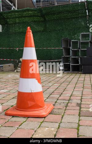Heller orangefarbener und weißer Verkehrskegel auf gepflastertem Boden in der Nähe eines Zauns als Sicherheitswarnung platziert. Stockfoto