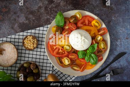 Oberblick auf einen frischen Tomaten- und Burrata-Salat mit bunten Tomaten, Basilikum, Oliven und Pinienkernen auf einem rustikalen karierten Tuch Stockfoto