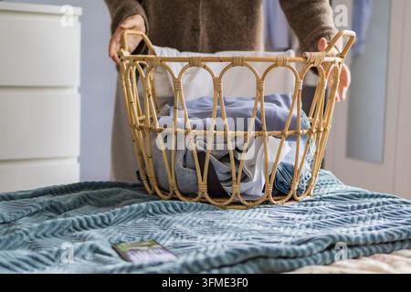 Mittlere erwachsene Frau hält einen Rattan-Wäschekorb voller Kleidung auf einem Bett mit blauer Decke. Konzept von Haushaltsorganisation, Hausarbeiten, Ordnung Stockfoto