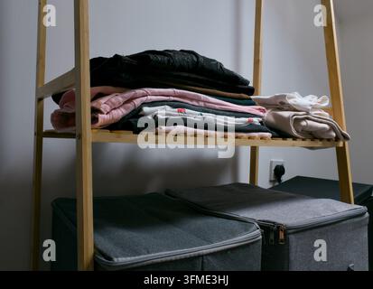 Eine Nahaufnahme zeigt ordentlich gefaltete Kleidung, die auf einem stilvollen Holzregal gestapelt ist, mit zwei grauen Aufbewahrungsboxen, die ordentlich auf dem Regal darunter platziert sind. Stockfoto