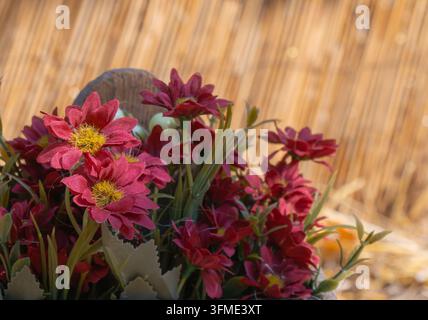 Nahaufnahme von roten künstlichen Stoffblumen mit gelben Mittelpunkten und grünen Blättern, angeordnet vor warmem Stroh für rustikales Deko- und Basteldesign. Stockfoto