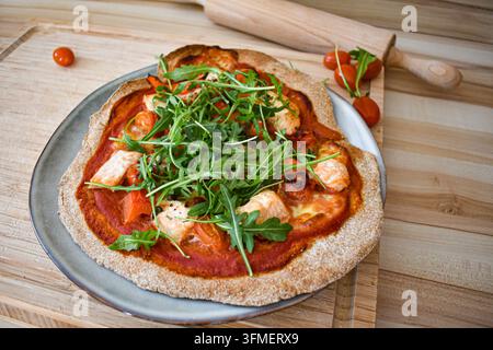 Frisch gebackene Sauerteigpizza mit Rucola, Tomaten und Lachs. Ein Nudelholz im Hintergrund und kleine frische Cocktailtomaten Stockfoto
