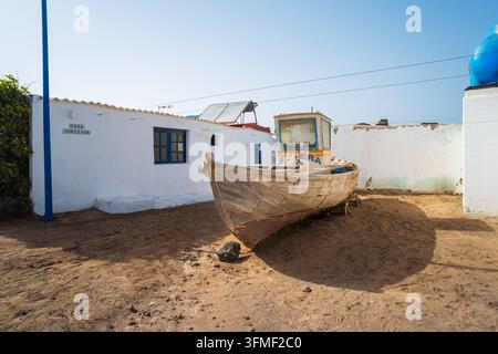 MAJANICHO, FUERTEVENTURA – 3. AUGUST 2026: Ein verwittertes hölzernes Fischerboot ruht auf dem Sand neben einem weiß getünchten Haus in Majanicho. Stockfoto