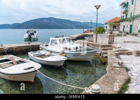 Blick von einem Betondock aus, der ein kleines weißes und blaues Motorboot zeigt, das in ruhigen Buchtgewässern vor Anker liegt, mit Blick auf Inseln und felsiger Küste in der Nähe von Przno und Sve Stockfoto