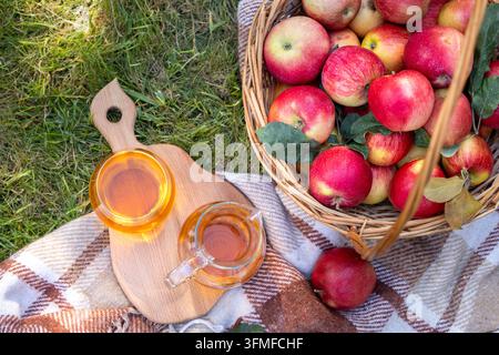 Körbe aus Weidengeflecht, der überquillt mit reifen roten und gelben Äpfeln auf einer karierten Decke. Neben einem Holzbrett mit einem Glas und einer Kanne, gefüllt mit Erfrischungen Stockfoto