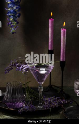 Zwei lila Lavendelbier-Cocktails in Martinigläsern mit Eis und frischen Lavendelstielen, umgeben von brennenden lila Kerzen und Blumen auf einem dunklen Tablett Stockfoto