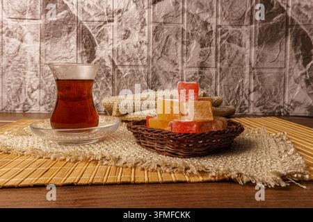 Komposition mit türkischem Tee in einer gläsernen 'Armudu'-Tasse und einem Haufen farbenfrohem Lokum in einem geflochtenen Korb. Serviert auf einer Bambusmatte und rauem Stoff agai Stockfoto