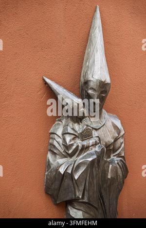 Metallenes Skulpturenrelief einer bußfertigen Figur in einer spitzen Kapuze vor einer rustikalen, strukturierten Wand, das die Traditionen der Karwoche in Zamora, Spanien, ehrt. Stockfoto
