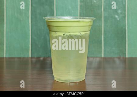 Eisgekühltes Matcha-Kokosnuss-Getränk in einem klaren Plastikbecher vor einer grün gekachelten Wand auf dunklem Holztisch Stockfoto