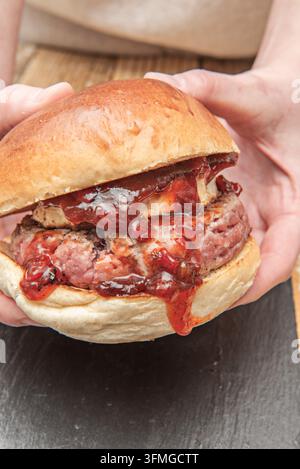 Nahaufnahme von zwei Händen, die einen dicken Burger aufziehen und einen saftigen Patty zeigen, der in zäher roter Sauce ertränkt ist Stockfoto