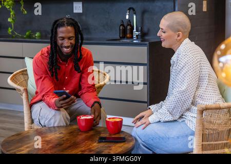 Verschiedene Freunde plaudern und trinken Kaffee am Kochnischentisch mit roten Tassen und Smartphones. Menschen, rund, Lounge, modern, gemütlich, Latte, Pflanze Stockfoto
