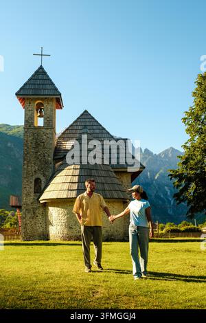 In Teth, Albanien, geht ein Paar an einem sonnigen Tag Hand in Hand, umgeben von atemberaubenden Berglandschaften. Die charmante Kirche bietet eine malerische Kulisse für ihre freudige Erkundung. Stockfoto