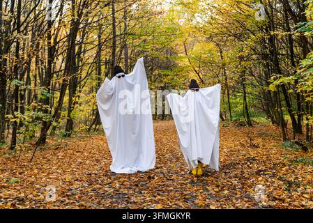 Zwei Personen in Halloween-Geisterkostümen stehen oder spazieren durch einen Herbstwald, umgeben von bunten Laub. Die Kostüme bestehen aus weißen Stofflaken, die die Figuren bedecken, und schaffen so eine verspielte und geheimnisvolle saisonale Szene. Gefallene Blätter bedecken den Waldboden, während Baumstämme und Äste die Charaktere einrahmen. Stockfoto
