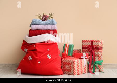 Weihnachtsgeschenke und Feiereinkäufe, ein Sack des Weihnachtsmanns gefüllt mit warmen Winterpullovern und verpackten Geschenken auf einem Holzboden in der Nähe einer beigen Wand Stockfoto