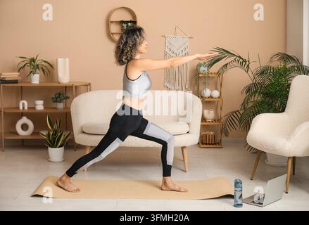 Wellness und Work-Life-Balance, Frau praktiziert Yoga zu Hause mit Online-Fitnesskurs auf dem Laptop in einem modernen Wohnzimmer-Interieur Stockfoto
