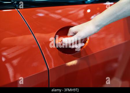 Nahaufnahme einer Person, die an einem modernen Autohaus in München, Deutschland, die Tür eines glänzenden roten Autos öffnet, und fängt die Vorfreude auf den Kauf eines neuen Fahrzeugs ein Stockfoto