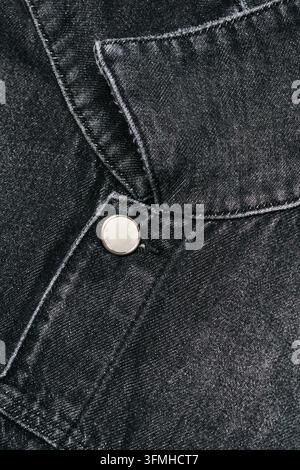 Nahaufnahme von strukturiertem schwarzem Denimstoff, genähten Nähten, gefaltetem Kleidungsdetail und einem runden Metallknopf. Stockfoto
