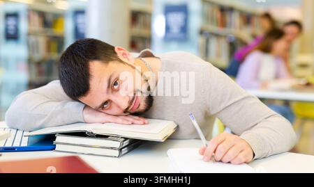 Fauler junger bärtiger Student, der auf Büchern am Tisch in der Bibliothek liegt Stockfoto