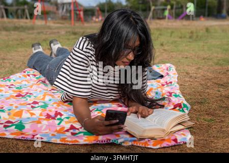 Frau liegt auf einer Picknickdecke im Park und hält ein Smartphone neben einem offenen Buch während einer ruhigen Pause im Freien. Stockfoto