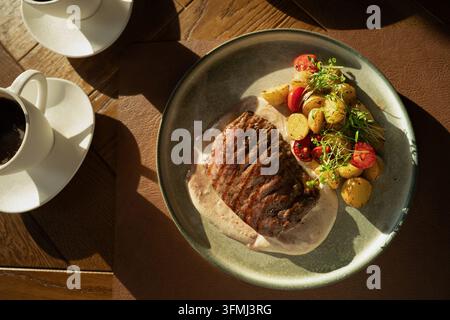 Gegrilltes Rindfleischsteak serviert mit cremiger Sauce, gebratenen Babykartoffeln, Kirschtomaten und frischen Kräutern auf einem Restauranttisch im warmen Sonnenlicht, repräsentiert Stockfoto